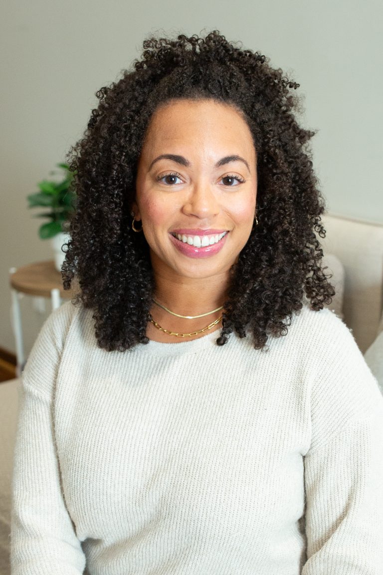 African American woman with brown curly hair sitting on a chair wearing a tan sweater