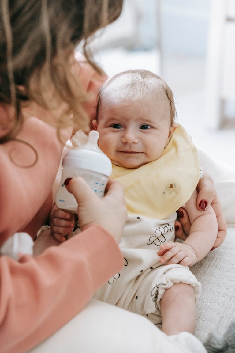Crop unrecognizable female with wavy hair in casual clothes hugging adorable newborn during feeding from bottle on sofa at home